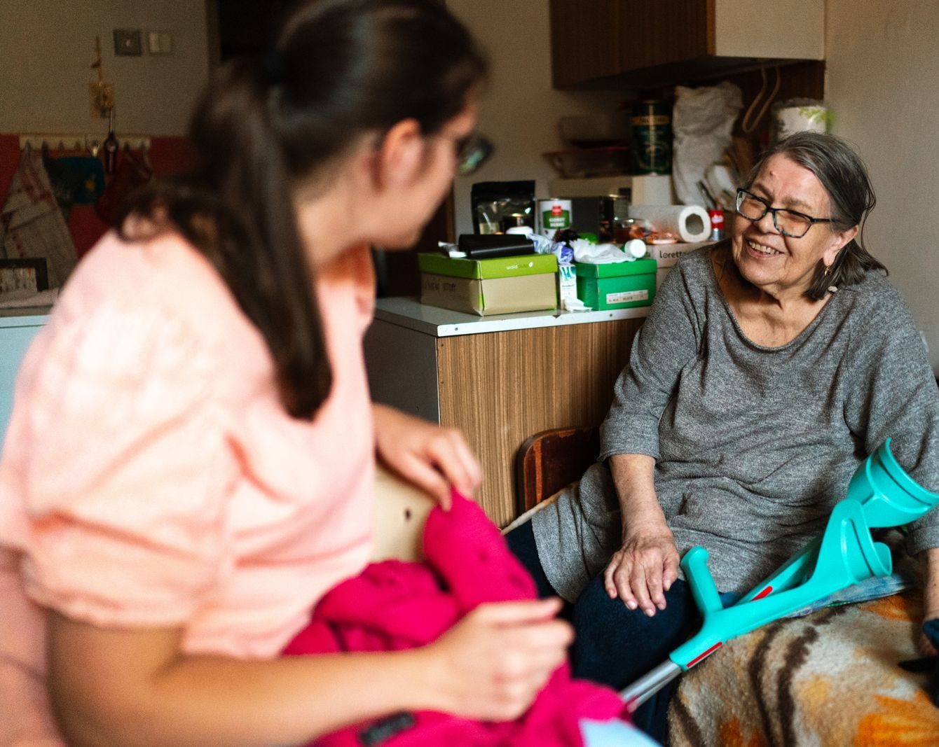 A caregiver helps a senior woman with a crutch in a cozy Prague home setting.